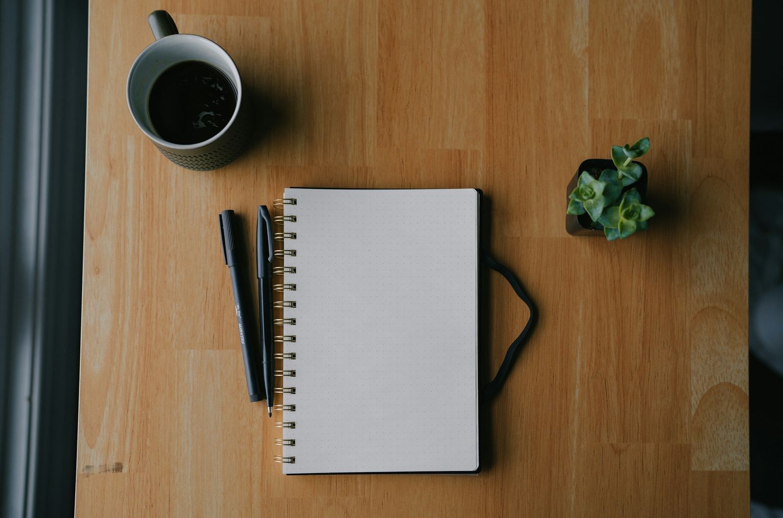 A blank notebook, coffee, and a small plant on a wooden desk